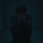 A person sitting alone in a dimly lit room, holding their head in their hands, looking overwhelmed and distressed. Shadows play across their face, emphasizing the emotional weight. The room is sparse, suggesting isolation, with muted, cool colors. The mood is somber and reflective, representing the burden of a secret, cinematic atmosphere, ultra realistic, highly detailed, 8k quality, photorealistic
