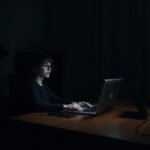 A quiet, dimly lit home office with a laptop screen glowing in the dark, a person sitting at a wooden desk with a look of stunned realization, soft shadows, moody atmosphere, realistic photography, high resolution.